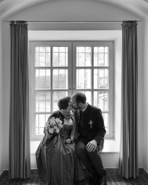 Brautpaar, dass sich in bayerischer Tracht an einem Fenster sitzend küsst